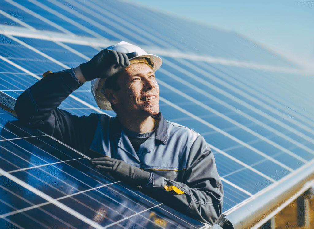 Solenergi med trygg installation – Åsa Elkoncept Elektriker med skyddshjälm arbetar vid solpaneler och blickar upp mot solen med ett leende.